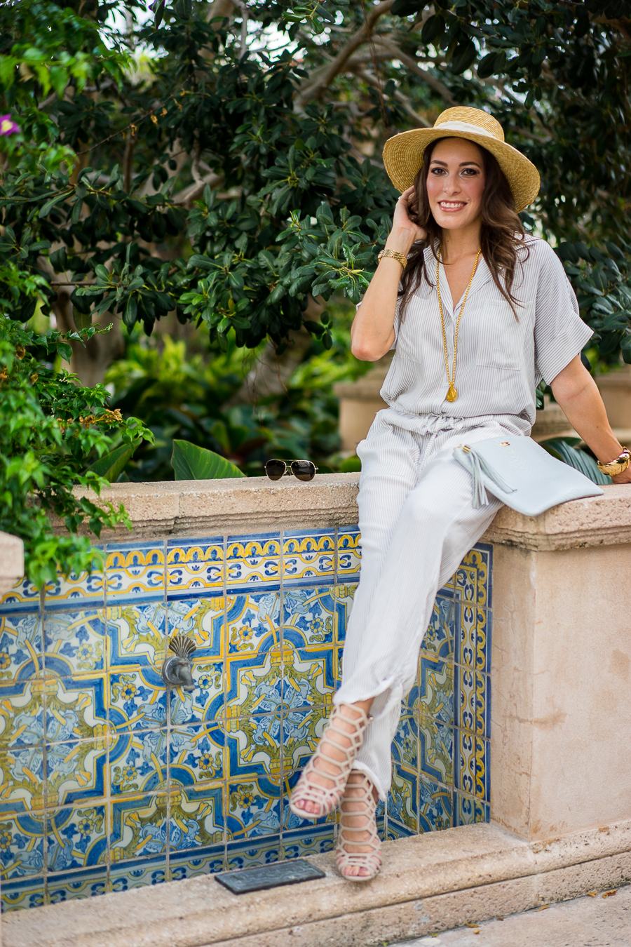 A Glam Lifestyle Blogger Amanda Champion Faithfull the Brand Striped Jumpsuit, with gold accessories and straw hat and holding her Gigi New York Uber clutch
