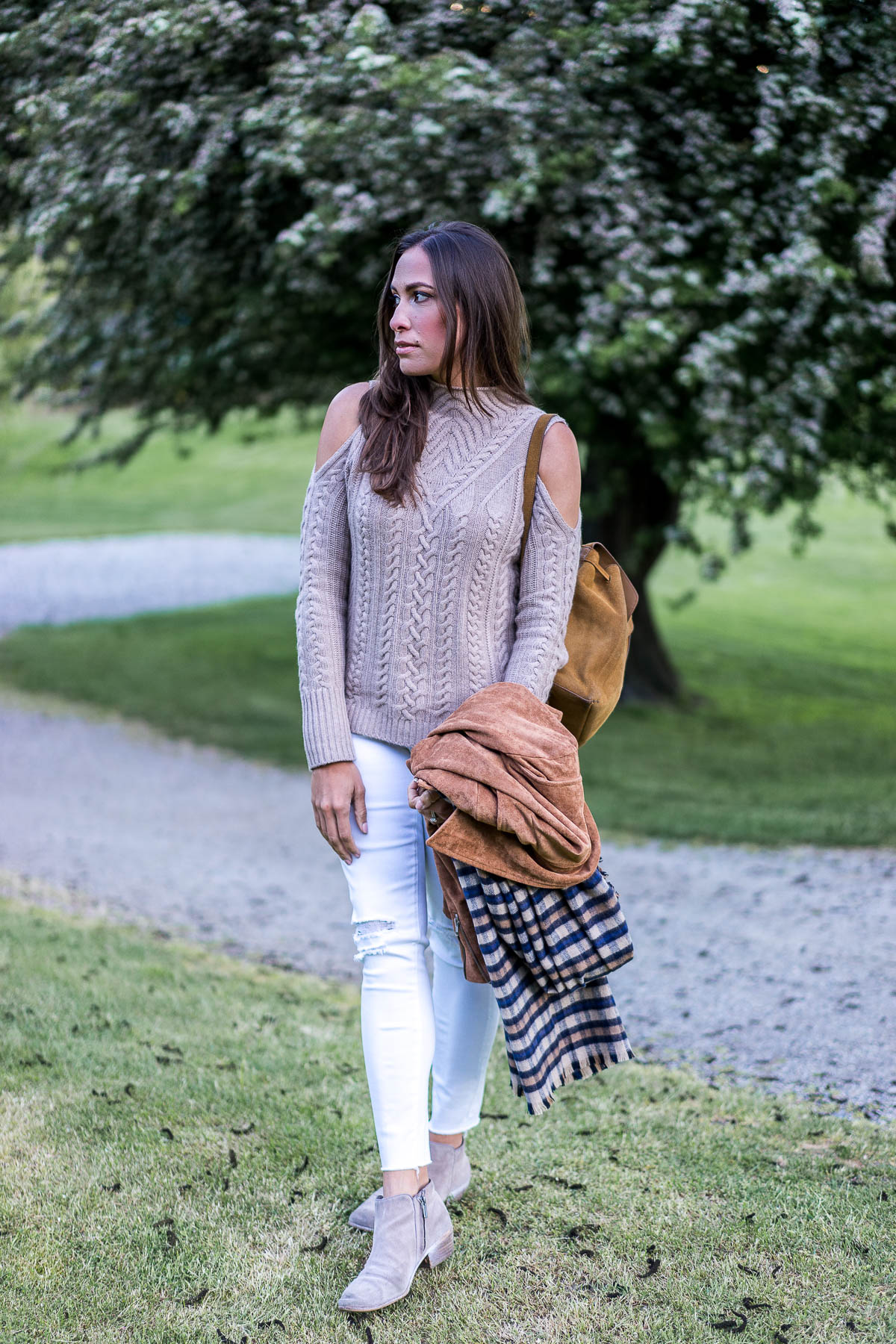 a glam lifestyle south florida fashion blogger carries tan suede moto jacket and plaid blanket scarf while wearing cashmere cold shoulder sweater and white legging jeans
