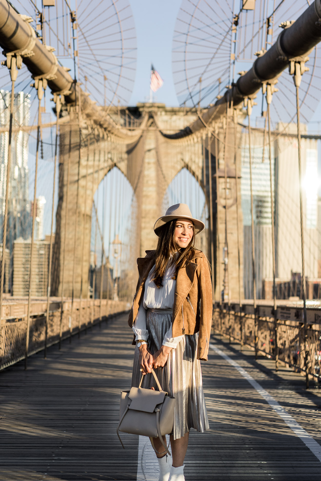 A metallic pleated skirt is a staple piece for Fall styled by Amanda of A Glam Lifestyle with her Celine belt bag, Topshop white booties and a Lulus camel suede jacket at the Brooklyn Bridge