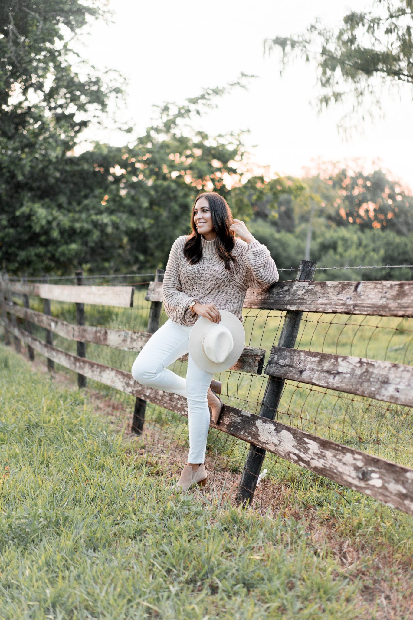 woman wearing hat, jeans, sweater leaning on a fence